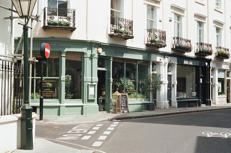 A street scene outside a shopfront with large glass windows displaying plants and decorative items, painted in a pastel green colour, with black signage above the window. The shop is located in a traditional British-style building with white walls and multiple upper-floor windows with black wrought-iron balconies. In front of the shop, a chalkboard sign indicates a menu or special offer, and several potted plants are positioned along the pavement. Nearby, a red and white no-entry traffic sign is mounted on a pole, while a nearby pavement has white dashed markings indicating a pedestrian or bicycle crossing. The environment is well-lit, suggesting daytime, and the area appears quiet with no pedestrians or vehicles visible. The setting reflects a typical urban street scene suitable for a home relocation or furniture transport service, with West Kensington Man and Van specialising in removals, including packing and loading processes.