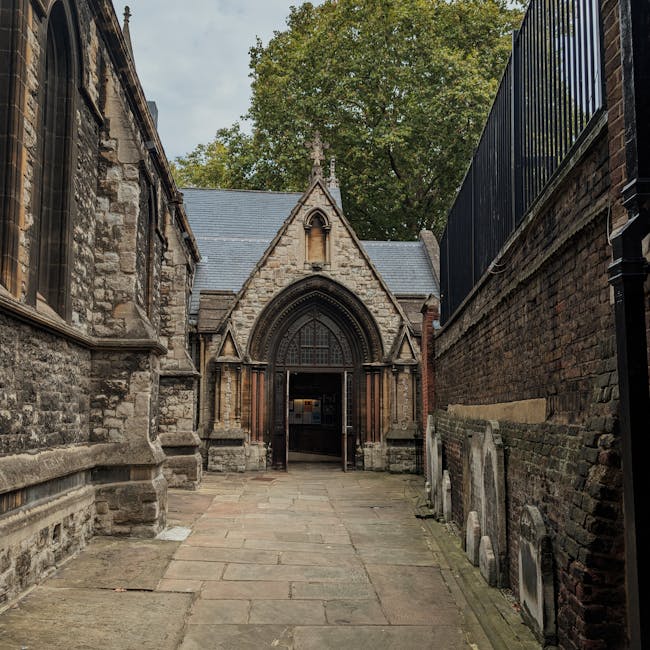 A narrow stone-paved pathway leading to an aged church entrance constructed from grey stone with a pointed gabled roof, situated between two brick walls. On the right side, a black metal fence runs along the top of a brick wall, while on the left, the church wall features windows with dark frames. In the foreground, several wheeled trolleys and packing materials such as cardboard boxes and plastic wrapping are visible, indicating active furniture transport or a home relocation process. The scene is outdoors under overcast skies, with surrounding greenery visible behind the church, suggesting a peaceful, residential or historic area. The overall setting implies an ongoing moving or packing operation supported by a professional removals company, highlighting aspects of furniture loading, packing, and site access in the context of house removals or estate clearance.