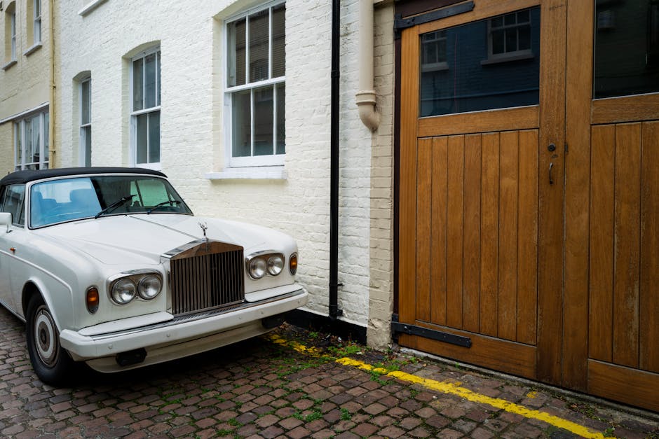 A vintage white luxury car with a black roof is parked on a cobblestone driveway next to a white brick building with four large double-hung sash windows. The car's front grille and headlights are prominent, and the car appears to be stationary. To the right, there is a large wooden sliding door with glass panels at the top, set into the building's exterior. The driveway is bordered by yellow paint markings indicating parking restrictions or loading zones. The surrounding environment includes a black downspout attached to the building, and the area is well-lit, suggesting daytime. This scene reflects a typical home relocation or furniture transport setting, with the vehicle prepared for loading or unloading. West Kensington Man and Van may assist with similar house removals or packing and moving services, particularly in areas with parking permit regulations like Hammersmith and Fulham.