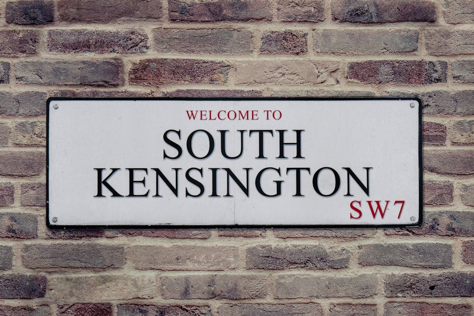 A rectangular white sign mounted on a brick wall, displaying the text 'WELCOME TO SOUTH KENSINGTON SW7' in black and red lettering. The sign is positioned at eye level and is secured with four bolts, one in each corner. The brick wall behind the sign consists of reddish-brown bricks with visible mortar joints. The lighting is natural, evenly illuminating the sign and wall. This image may relate to house removals or house relocation services, as part of the process of moving goods from or within the South Kensington area, with West Kensington Man and Van providing professional removals and transport solutions.