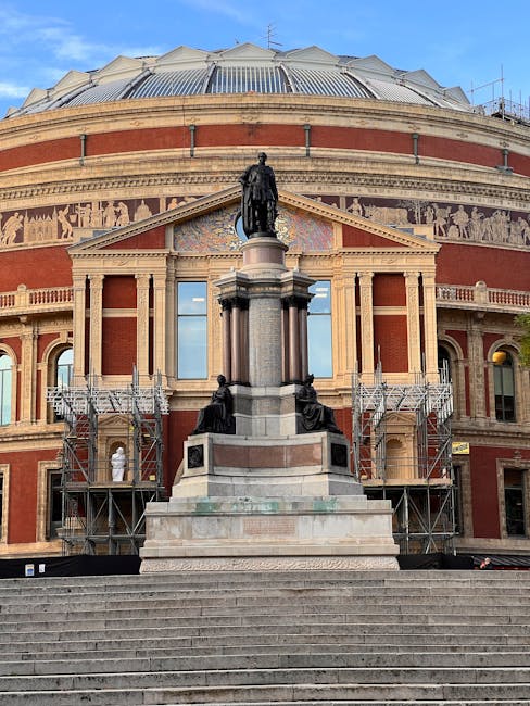 The exterior of a historic building with a domed roof and a large, rounded façade constructed of red brick and light stone detailing. In front of the building, there is a prominent statue of a standing figure atop a tall pedestal, flanked by two seated statues at the base. The pedestal is situated on a broad, stepped platform accessed by a set of stone stairs leading up from the pavement. Scaffolding surrounds parts of the building, indicating ongoing maintenance or restoration work, and the scene is illuminated by natural daylight with a clear sky overhead. The image captures the detailed architecture, the statues, and the construction elements, reflecting the craftsmanship and ongoing preservation efforts typical in historic London landmarks, relevant to house removals or relocation services involved in moving or restoring valuable or historic properties, as offered by West Kensington Man and Van.