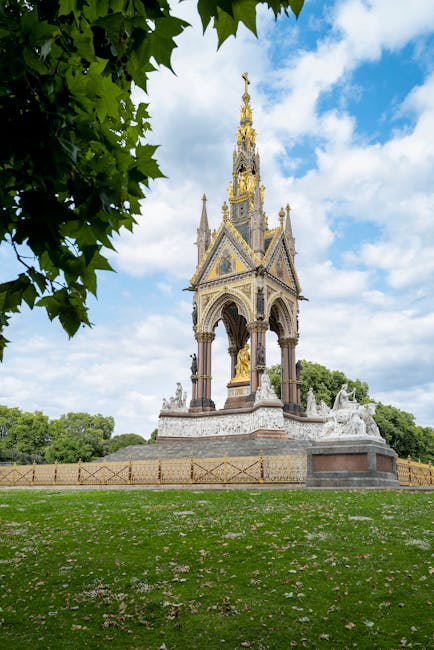 The image shows an ornate, Victorian-style monument situated on a circular, paved area within a green park, with well-maintained grass and scattered fallen blossom petals. The monument features intricate stone and metalwork, including tall, slender spires topped with decorative finials, and gilded accents highlighting the structure's details. Surrounding the base are sculptural elements and small statues, with a protective wooden fence enclosing the installation. In the foreground, a large leafy tree with vibrant green leaves partially frames the scene on the left side, while the background reveals a partly cloudy sky with patches of blue and white clouds, and lush trees lining the horizon. The setting is outdoors, capturing natural daylight that illuminates the monument and the park area, representing a typical urban greenspace that could be part of a house removals or relocation process, with West Kensington Man and Van potentially providing services related to moving or clearance in the vicinity.
