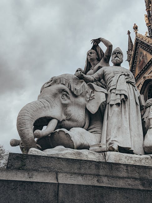 The exterior of a historic building with a domed roof and a large, rounded façade constructed of red brick and light stone detailing. In front of the building, there is a prominent statue of a standing figure atop a tall pedestal, flanked by two seated statues at the base. The pedestal is situated on a broad, stepped platform accessed by a set of stone stairs leading up from the pavement. Scaffolding surrounds parts of the building, indicating ongoing maintenance or restoration work, and the scene is illuminated by natural daylight with a clear sky overhead. The image captures the detailed architecture, the statues, and the construction elements, reflecting the craftsmanship and ongoing preservation efforts typical in historic London landmarks, relevant to house removals or relocation services involved in moving or restoring valuable or historic properties, as offered by West Kensington Man and Van.