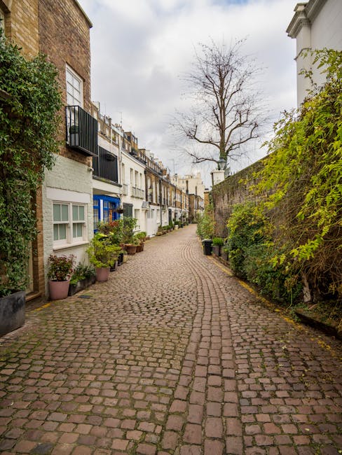 A narrow cobbled street in West Kensington with rows of terraced houses on both sides, featuring white and pastel-colored facades, some with small balconies and window boxes. Potted plants are arranged along the pavement near the entrances. A large, leafless tree stands at the end of the street under an overcast sky. The scene illustrates typical residential architecture in an area where house removals or local moving services might encounter tight access challenges, as discussed on the page about narrow access removals at West Kensington common problems by West Kensington Man and Van.