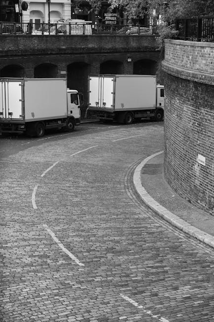 A narrow cobbled driveway with a curved path, leading to a parking area with three white moving trucks parked parallel to a brick wall and an arched bridge. The trucks are positioned for a home relocation or furniture transport, possibly part of a house removal service. The area is bordered by a metal railing above the bridge, with trees and residential buildings visible in the background. The setting appears to be an urban environment with limited access, highlighting potential challenges for large vehicle manoeuvres during a move. The scene is in black and white, with natural lighting emphasizing the textures of the cobblestones and brickwork. West Kensington Man and Van may be involved in coordinating transport and loading operations in this confined space, focusing on packing, moving, and logistics within a restricted access area.