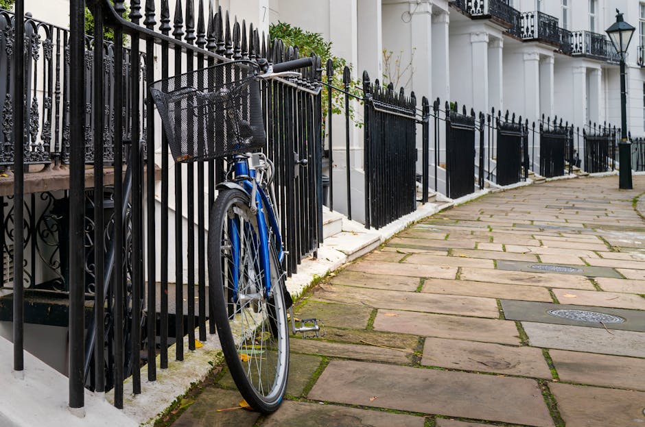 A narrow cobbled street in West Kensington with rows of terraced houses on both sides, featuring white and pastel-colored facades, some with small balconies and window boxes. Potted plants are arranged along the pavement near the entrances. A large, leafless tree stands at the end of the street under an overcast sky. The scene illustrates typical residential architecture in an area where house removals or local moving services might encounter tight access challenges, as discussed on the page about narrow access removals at West Kensington common problems by West Kensington Man and Van.