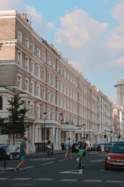 Image showing the exterior of a multi-storey residential building in a city environment, with several ground-floor entrances and large windows. The building is constructed of light-colored brick and has decorative architectural details along the façade. In front of the building, the street includes parked cars and a paved pavement. There are pedestrians walking on the sidewalk, including a person riding a bicycle across a crosswalk, and another person walking nearby. The scene is captured during daylight with a cloudy sky overhead. This setting illustrates a typical urban area suitable for home relocation and furniture transport, often serviced by professional removals companies like West Kensington Man and Van during packing and loading processes for house removals or short-distance moves.
