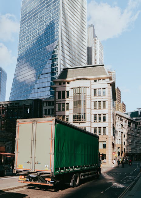 A large green cargo truck with a closed container is parked on a city street in front of modern glass skyscrapers and older stone buildings. The truck is positioned near the curb, with its rear doors visible and secured with metal latches. The street appears busy with some pedestrians and other vehicles in the background. The scene is set during daylight with clear skies and natural sunlight illuminating the buildings and the loading area. This image depicts a typical urban environment suitable for house removals and furniture transport services, as provided by West Kensington Man and Van, focusing on efficient loading and transport in a city setting.