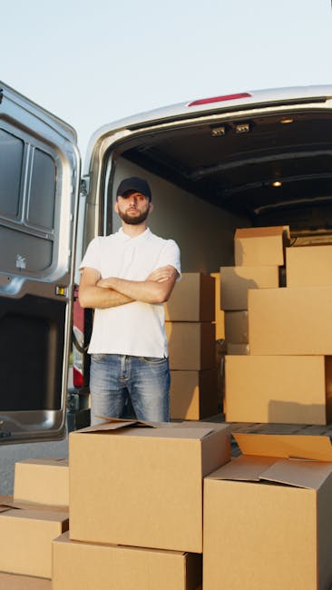 A man wearing a white t-shirt, jeans, and a dark cap stands with arms crossed in front of the open rear doors of a white moving van during a home relocation process. Inside the van, there are several neatly packed cardboard boxes of varying sizes, some stacked on top of each other, with a few placed on the pavement nearby. The boxes are secured with plastic wrap and are ready for transport, indicating an organized packing and loading process. The scene takes place outdoors under bright, natural daylight, with the van positioned on a paved driveway or street. The background includes part of the building or surrounding area, but the focus remains on the man, the boxes, and the vehicle, illustrating professional furniture transport and moving logistics services as offered by West Kensington Man and Van, supporting efficient house removals and packing services.