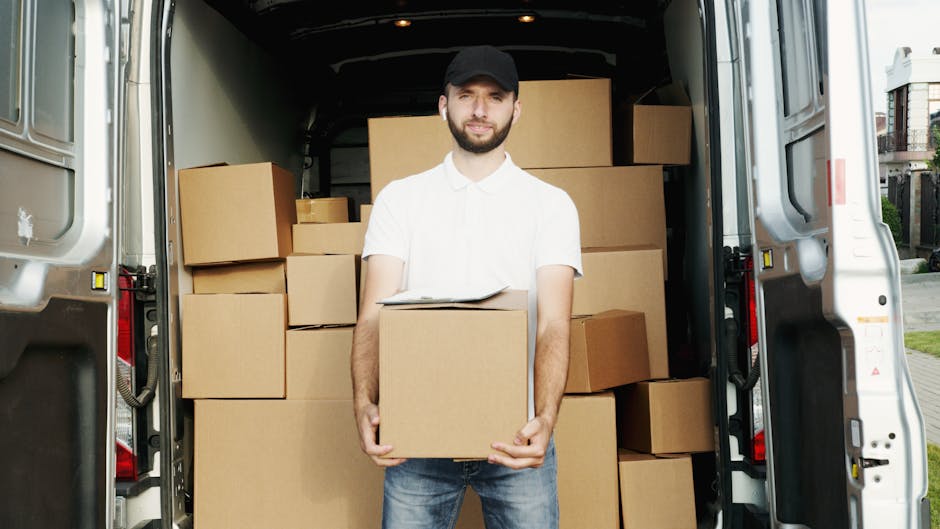 A man wearing a white polo shirt, dark jeans, and a black cap stands inside the open rear of a white moving van parked outdoors, holding a cardboard box in front of him. The van's interior is filled with stacked cardboard boxes of various sizes, some wrapped in plastic or placed on top of others, indicating a house removal or furniture transport process. The scene takes place on a paved area near residential buildings, with natural daylight illuminating the scene, highlighting the loading process involved in home relocation services offered by West Kensington Man and Van. The image captures the careful handling of packing materials and the logistical aspects of packing and moving procedures, aligning with professional removals service offerings in the West Kensington area.