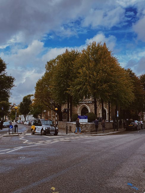 A view of a street outside a historic building with a rounded arch entrance, partially obscured by large trees with green and brown foliage, indicating an autumn setting. Several pedestrians are walking on the pavement, and a few vehicles are parked or moving along the road. In the foreground, a small van is parked near a crossing with tactile paving and bollards. The scene depicts a typical urban environment, likely during daytime with partly cloudy skies. This image relates to house removals and home relocation services offered by West Kensington Man and Van, illustrating the logistical environment in which furniture transport and packing activities take place.