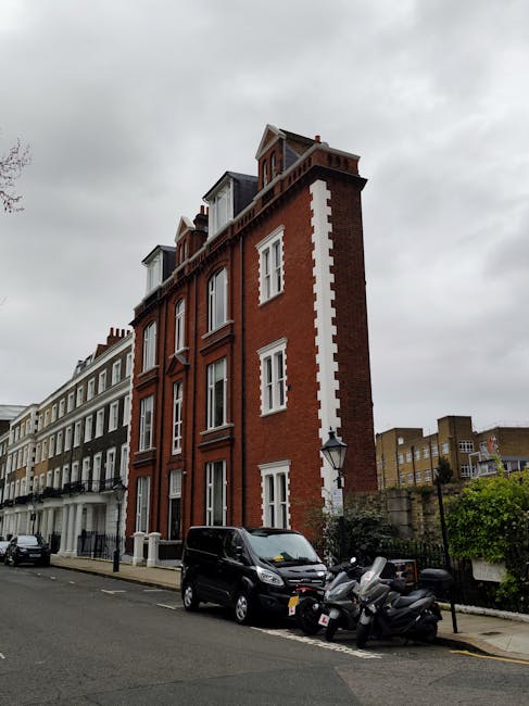 A multi-storey red brick building with white window frames and decorative corner stones, situated on a city street under an overcast sky. The building has several floors with large sash windows, and the uppermost floor features dormer windows in the roof. In front of the building, on the pavement, there are three parked vehicles: a black van, a dark grey car, and a group of three motorcycles. The area is part of a residential street, with adjacent buildings visible to the left, and some greenery and a lamppost near the sidewalk. The photograph captures the scene during daylight hours, with diffused natural light due to the cloudy sky, illustrating an urban environment suitable for house removals and furniture transport services as coordinated by companies like West Kensington Man and Van.